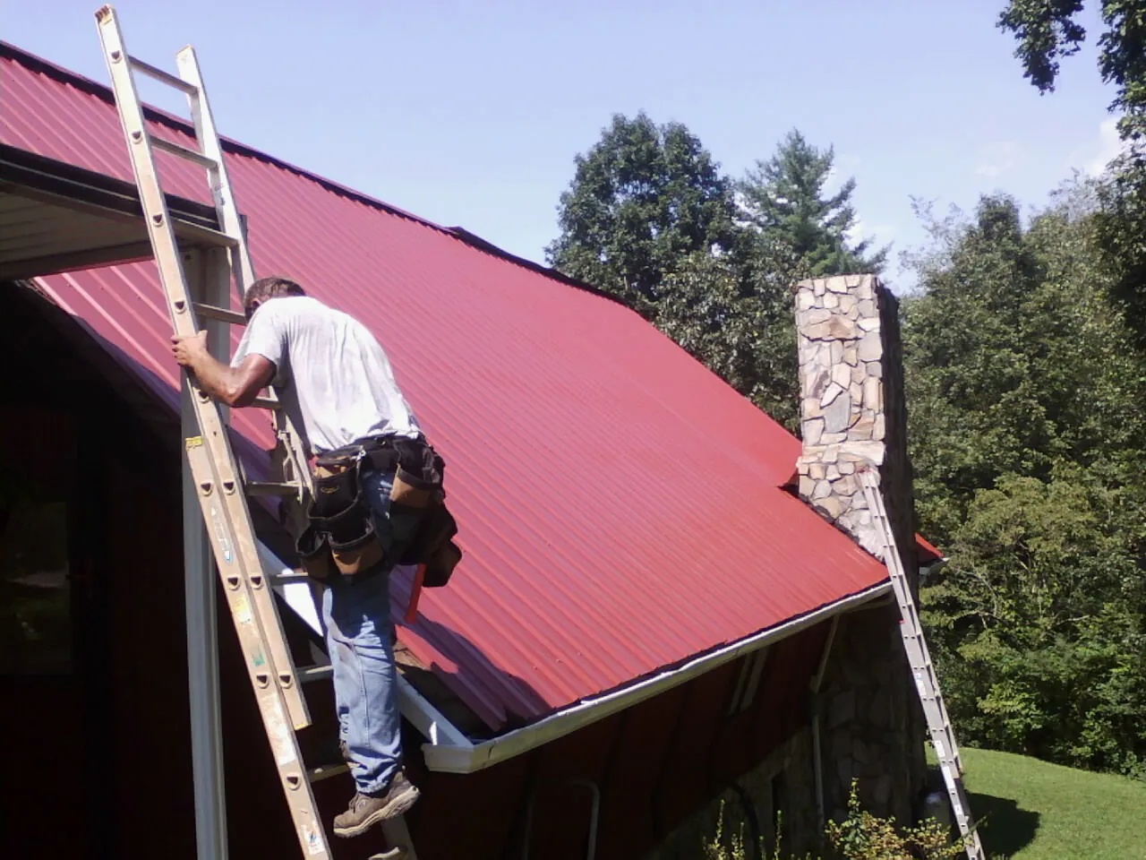 Roofer on a ladder beside a red metal roof and stone chimney — Soffit & Fascia Repair by Roofing Brothers in Hooks